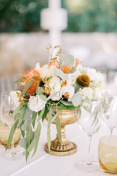 Colorful bouquet of flowers in a bronze vase stands among glasses on a served table. High quality photo