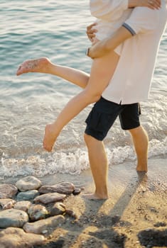 Man holds in his arms woman waving her legs while standing on the seashore. Cropped. Faceless. High quality photo