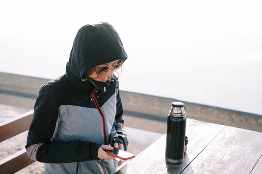 Traveler in a jacket with a pulled hood sits at a table and looks at a smartphone. High quality photo