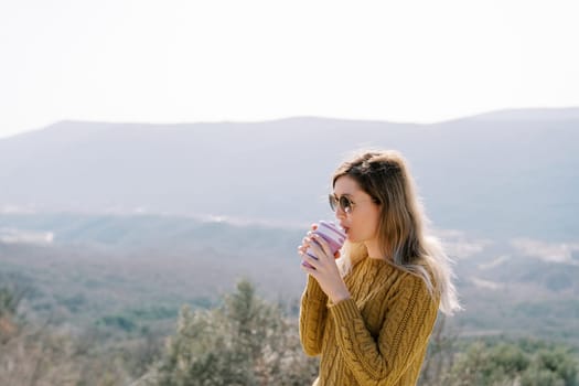 Young woman in sunglasses drinking coffee from a glass while standing in the mountains. High quality photo