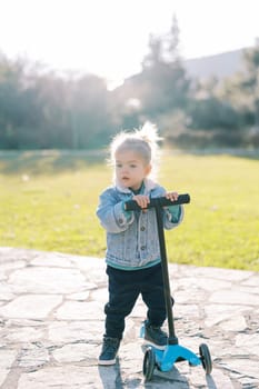 Little girl stands with a scooter on a path in the park. High quality photo