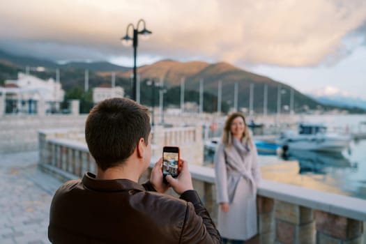 Man takes a picture with his phone of woman at the fence on the pier with moored boats. Back view. High quality photo