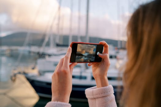 Woman shoots a marina with yachts at sunset with a smartphone. Back view. High quality photo