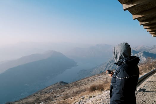 Man in a hooded jacket with a thermos mug stands in the mountains and looks at the bay. Side view. High quality photo