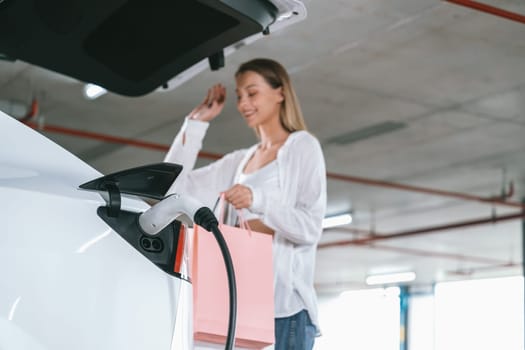 Young woman travel with EV electric car to shopping center parking lot charging in downtown city showing urban sustainability lifestyle by green clean rechargeable energy of electric vehicle innards