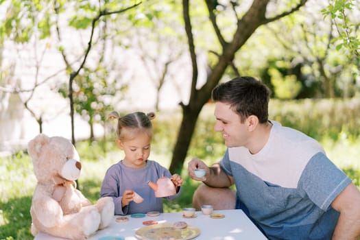 Smiling dad with a cup in his hand looks at a little girl pouring tea from a toy teapot. High quality photo