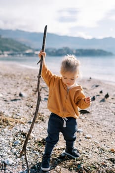 Little girl with a stick in her hand walks along the pebble beach looking at her feet. High quality photo