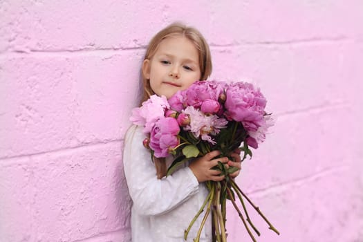 Portrait of a little girl with the peonies near pink wall