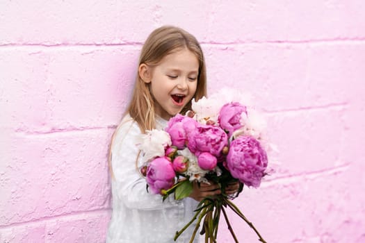 Portrait of a little girl with the peonies near pink wall