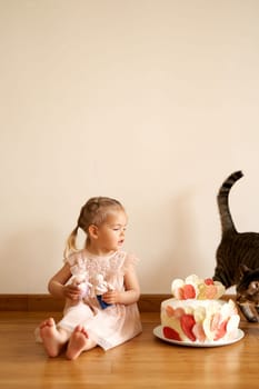 Little girl sits on the floor and looks at a cat sniffing a birthday cake on a plate. High quality photo