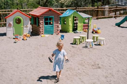 Little girl walks barefoot on the sand on the playground with sandals in her hands. High quality photo