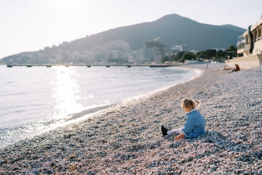 Little girl is sitting on a pebbly beach by the sparkling sea. Side view. High quality photo