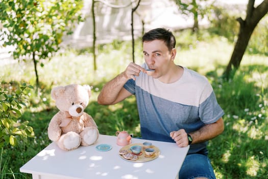 Dad drinks tea from a toy cup at the table with a teddy bear at a tea party in the garden. High quality photo