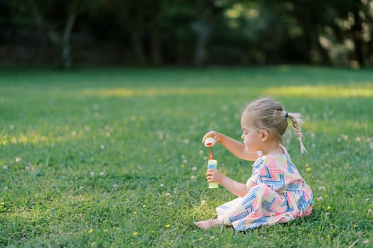 Little girl is sitting on a green meadow with a bottle of soap bubbles in her hands. Side view. High quality photo