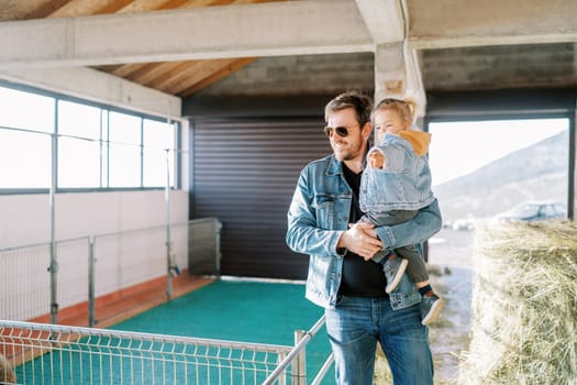 Little girl pointing her finger at the corral while sitting in her dad arms on the farm. High quality photo