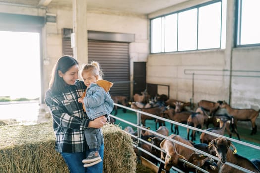 Mom with a little girl in her arms stands by a haystack near a goat paddock. High quality photo