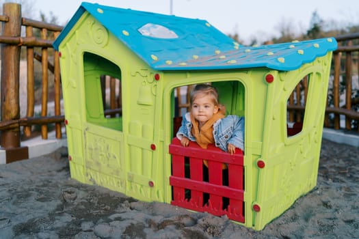 Little girl looks out the window of a toy house on the playground. High quality photo