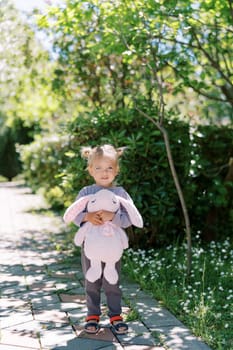 Smiling little girl with a stuffed bunny stands on a paved path in a sunny park. High quality photo