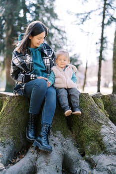 Smiling mother with a little girl are sitting on a stump covered with green moss in the forest. High quality photo