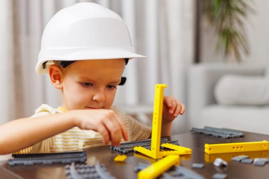 Child boy in helmet playing and building with colorful plastic bricks at the table.