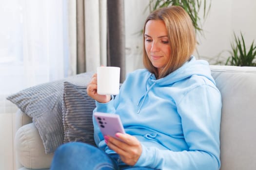 A woman unwinds on a comfortable sofa, smartphone in hand, and a cup of tea, embracing a perfect blend of tech and tranquility