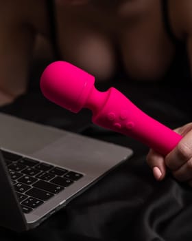 Woman holding pink vibrator next to laptop while lying on black sheet