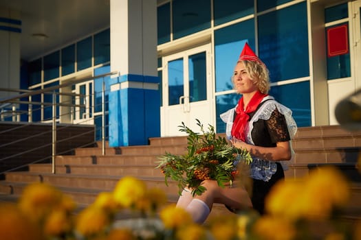 A Girl in black school uniform, white apron and red tie on steps of school with bouquet of flowers. Nostalgia photo shoot of teenager of female pioneer from USSR costume for September 1 or graduation