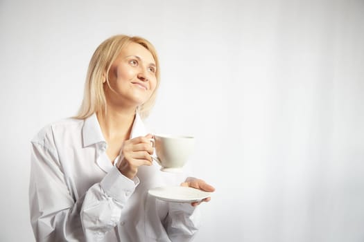 Portrait of pretty blonde smiling woman posing with cup of tea or coffee on white background. Happy girl model in white shirt in studio. The concept of pleasant morning at home or work. Copy space