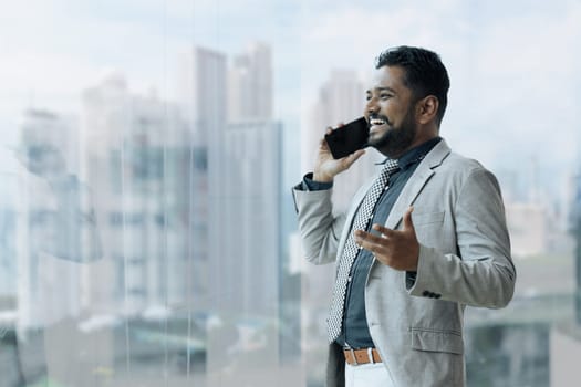 Indian businessman using smartphone talking having phone call conversation in office looking out window.