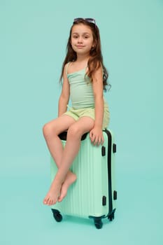 Vertical emotional portrait of a charismatic adorable little child girl, traveler going for summer holidays, sitting on her polyarbonate trendy suitcase, smiles at camera, isolated on blue background