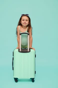 Caucasian happy child, beautiful little traveler girl with turquoise suitcase, smiles looking at camera, isolated on blue studio background. Kids. Travel. Weekend getaway and summer holidays concept