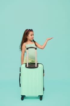 Caucasian pretty little traveler girl in summer wear, posing with turquoise suitcase, holding imaginary copy advertising space on her hand palm up, isolated over blue studio background. Kids and Trip