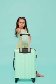 Caucasian happy traveler girl with turquoise suitcase, gestures with a thumb up, expressing agreement and satisfaction, looking at camera, isolated over blue studio background. Kids and travel concept