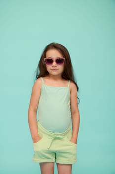 Charming elementary age child girl in summer clothes and sunglasses, putting hands in pockets, looking at camera, isolated blue background. Copy advertising space for text. Kids. Lifestyle. Emotions