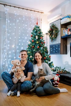 Smiling parents with a little girl and a cat on their knees sit near the Christmas tree on the floor. High quality photo