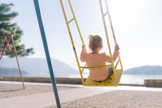 Little girl swings on a swing by the sea in bright sunlight. Back view. High quality photo