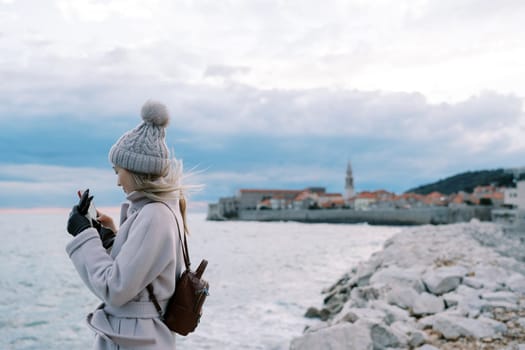 Girl with a backpack stands on the seashore and looks into her smartphone. Side view. High quality photo