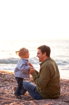 Little girl stands near a smiling dad sitting on the beach holding his hands. High quality photo