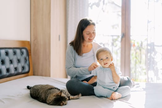 Mom sits with a little girl on the bed and combs her next to a lying tabby cat. High quality photo