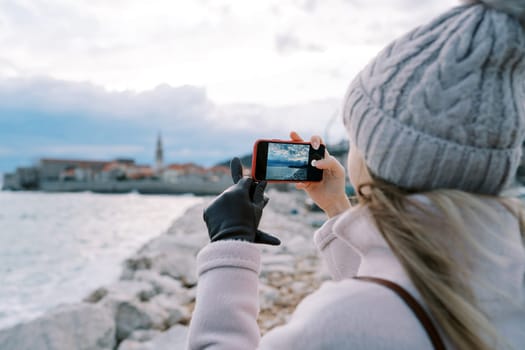 Young woman stands on the shore and shoots the sea with a smartphone. Back view. High quality photo