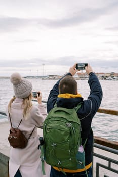 Man and woman shoot a port on the seashore with smartphones. Back view. High quality photo