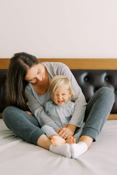 Smiling mom hugs a little laughing girl sitting behind her on the bed. High quality photo