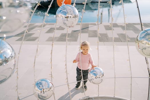 Little girl stretches her hands to a Christmas installation with mirror balls. High quality photo