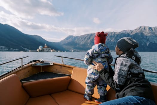 Mom sits in a boat holding a small child standing nearby and looks at the mountains. Back view. High quality photo