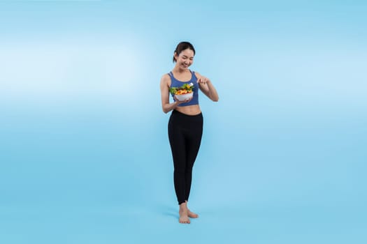 Young sporty Asian woman in sportswear holding salad bowl fill with vibrant of fruit and vegetable. Natural youthful and fit body lifestyle with balance nutrition on isolated background. Vigorous