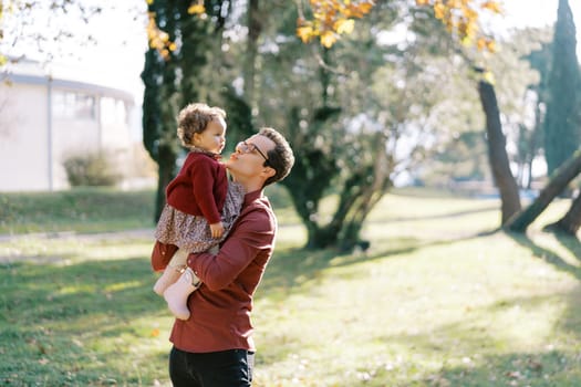 Dad pulls his lips to kiss a little girl in his arms while standing in the park. High quality photo