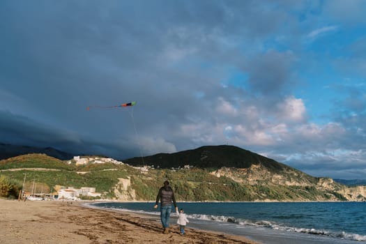 Dad and little daughter are walking along the seashore with a soaring kite. Back view. High quality photo