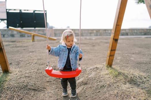 Little girl stands near the swing on the playground holding on to the ropes. High quality photo