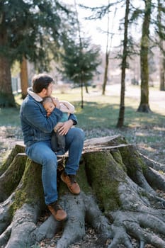 Dad hugs a little girl sitting on his lap on a big stump and looks into the forest. High quality photo