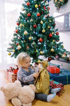 Little girl sits on the floor near the Christmas tree and unpacks a gift. High quality photo